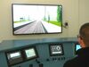Person sitzt vor einem Zug-Simulator mit mehreren Bildschirmen und steuert durch eine virtuelle Eisenbahnstrecke auf einem großen Bildschirm