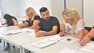 Vier schwarz, weiß und blau gekleidete Personen sitzen an einem Tisch in einem Klassenzimmer, konzentriert auf ihre Arbeit und umgeben von Büchern und Papieren.